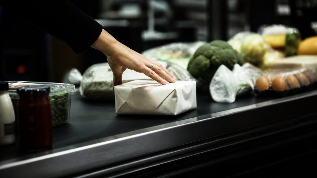 Supermarket checkout conveyor belt piled with grocery items, an anonymous hand reaching for a package, harsh fluorescent light overhead