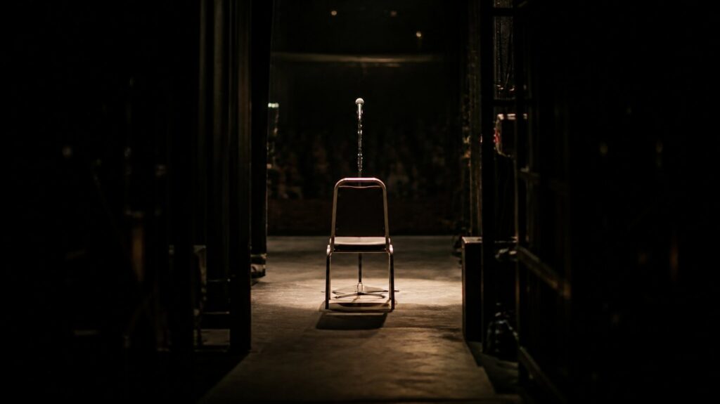 Crowded backstage corridor with a single empty spotlit chair facing a dark auditorium, microphone stand waiting under warm light
