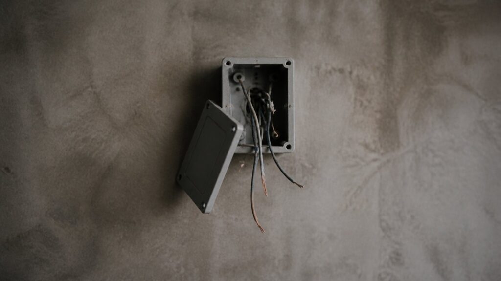 A grey electrical junction box on a bare wall, its cover half open, loose disconnected wires hanging limply in dull overcast daylight.