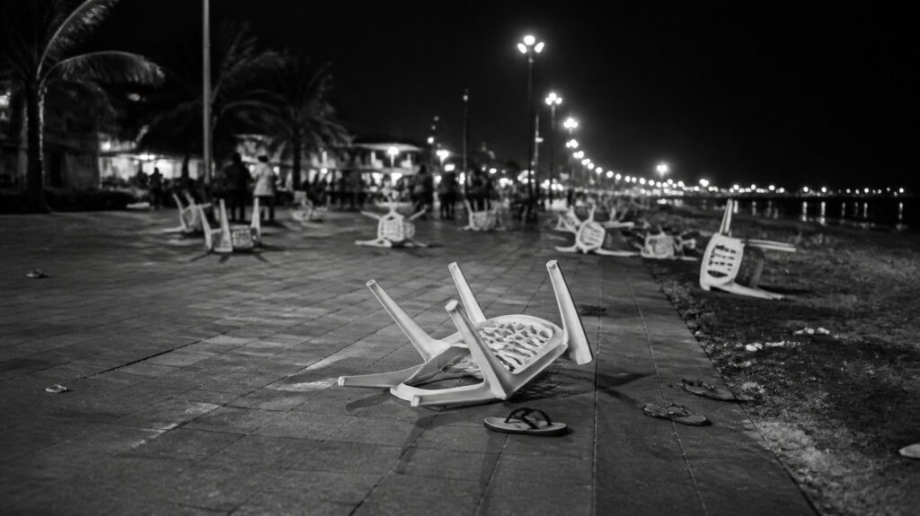 Crowded seaside boardwalk at night, empty overturned plastic chairs and scattered sandals on the pavement, streetlights glowing overhead